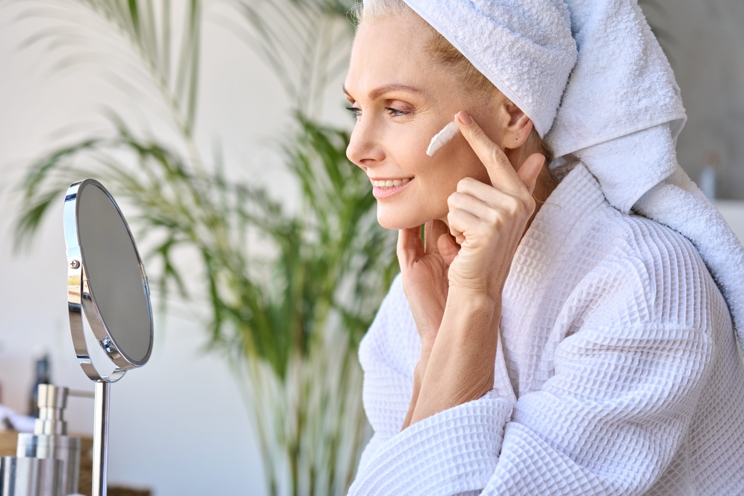 Gorgeous senior older adult 50 years old blonde woman wearing bathrobe and turban towel in bathroom applying moisturizing tightening face skin treatment, looking at mirror. Morning beauty routine.