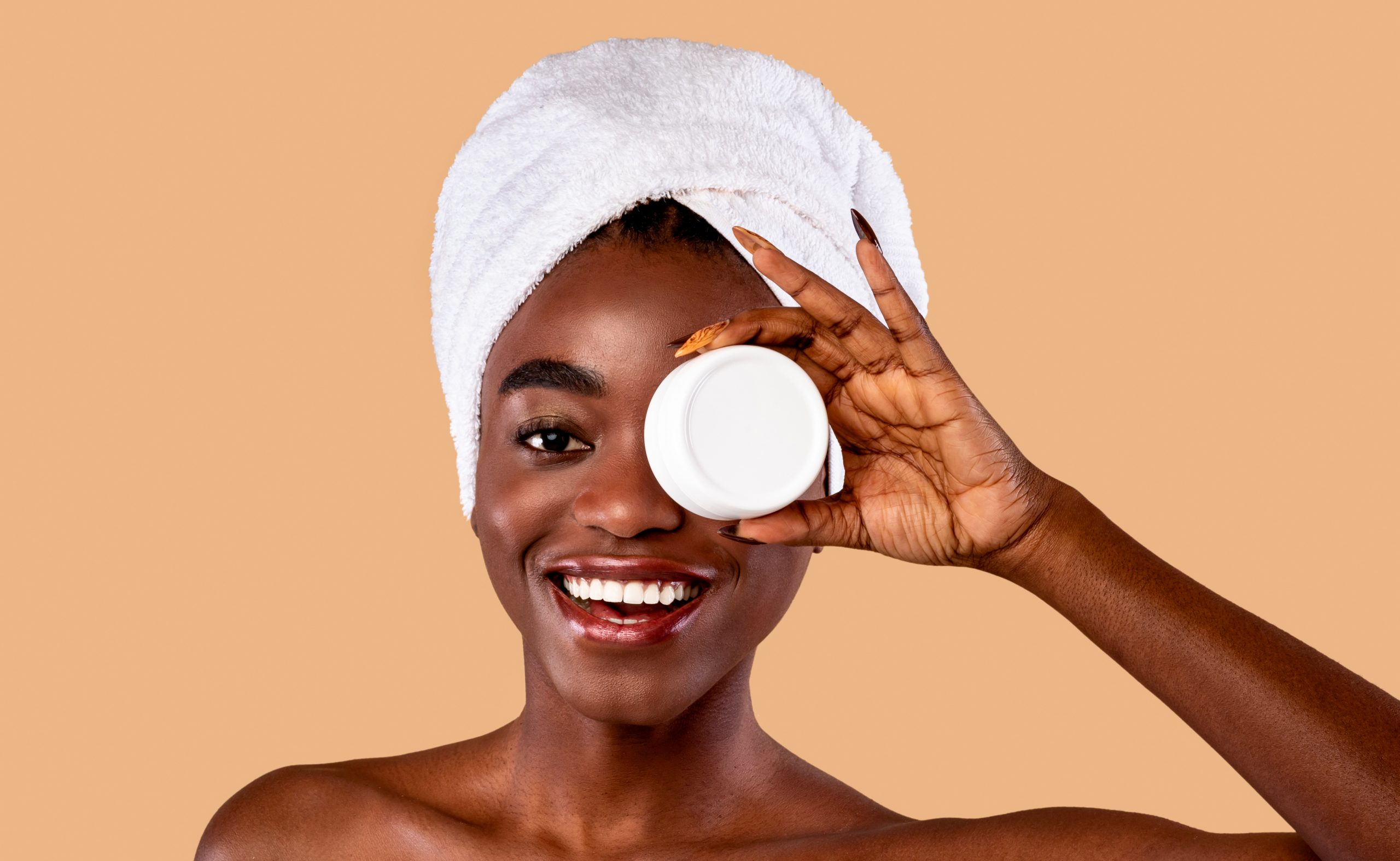 Skin Care. Beauty Portrait Of Happy Black Woman Holding White Round Jar With Moisturising Cream In Hand, Covering One Eye With New Skincare Product, Standing Wrapped In Towel Over Studio Background