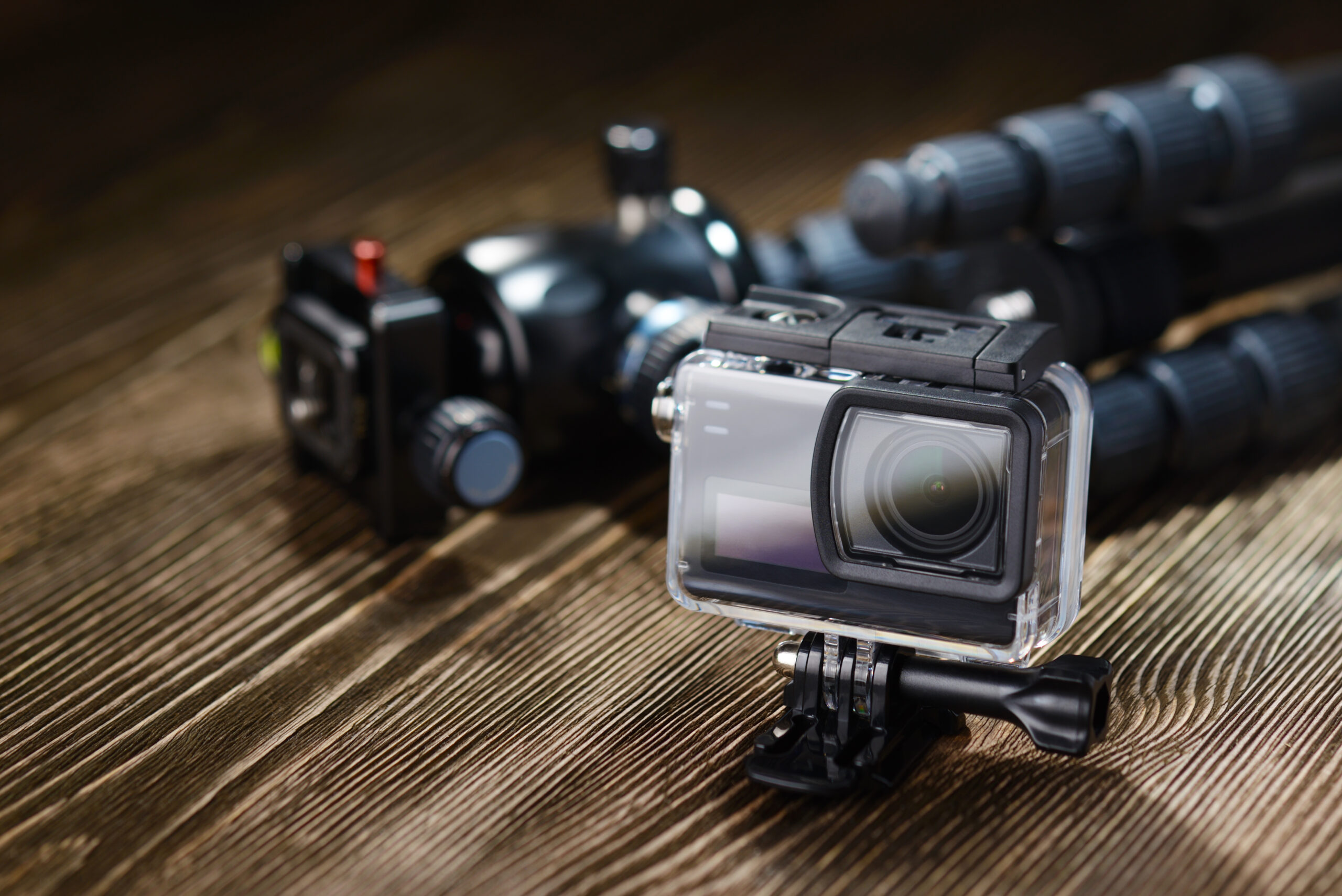 Black small action camera in waterproof housing and tripod on brown wooden tabletop.