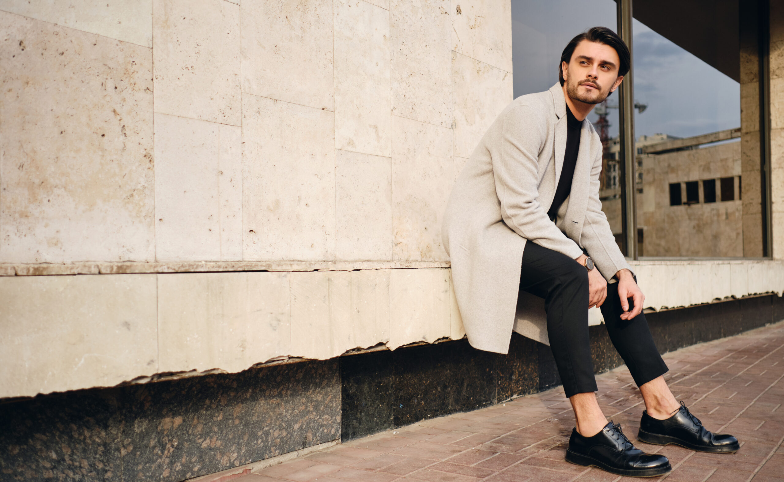 Young handsome casual man in coat thoughtfully looking away sitting on street