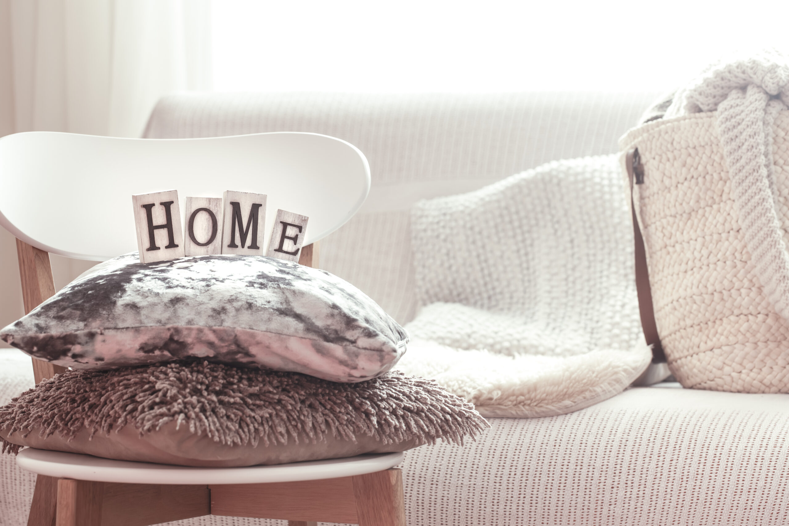 Home decoration. Decorative pillows and home lettering on white wooden stylish chair. Sofa and wicker basket with cushions in the background