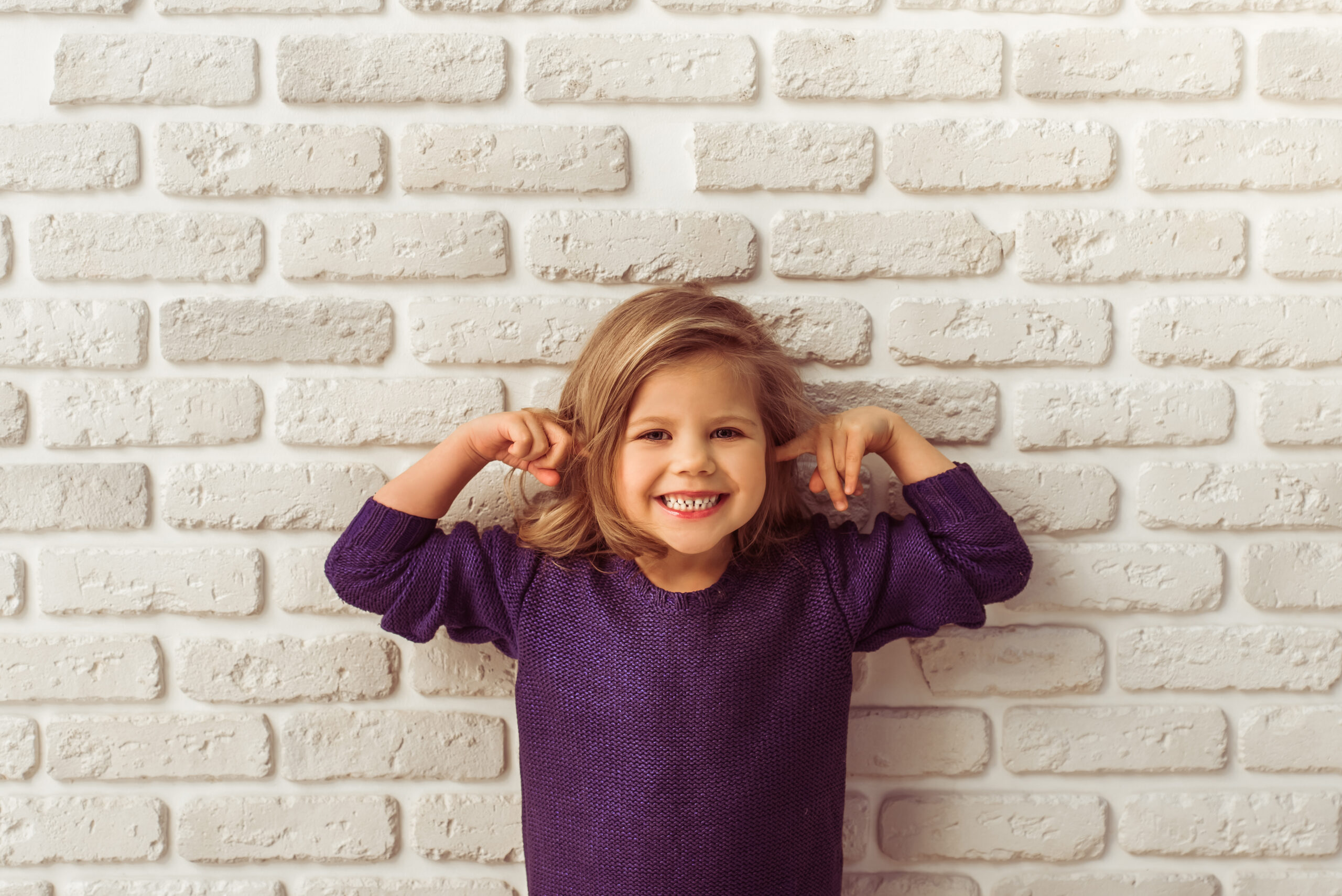 Beautiful little girl in casual clothes is covering her ears, looking at camera and smiling, standing against white brick wall