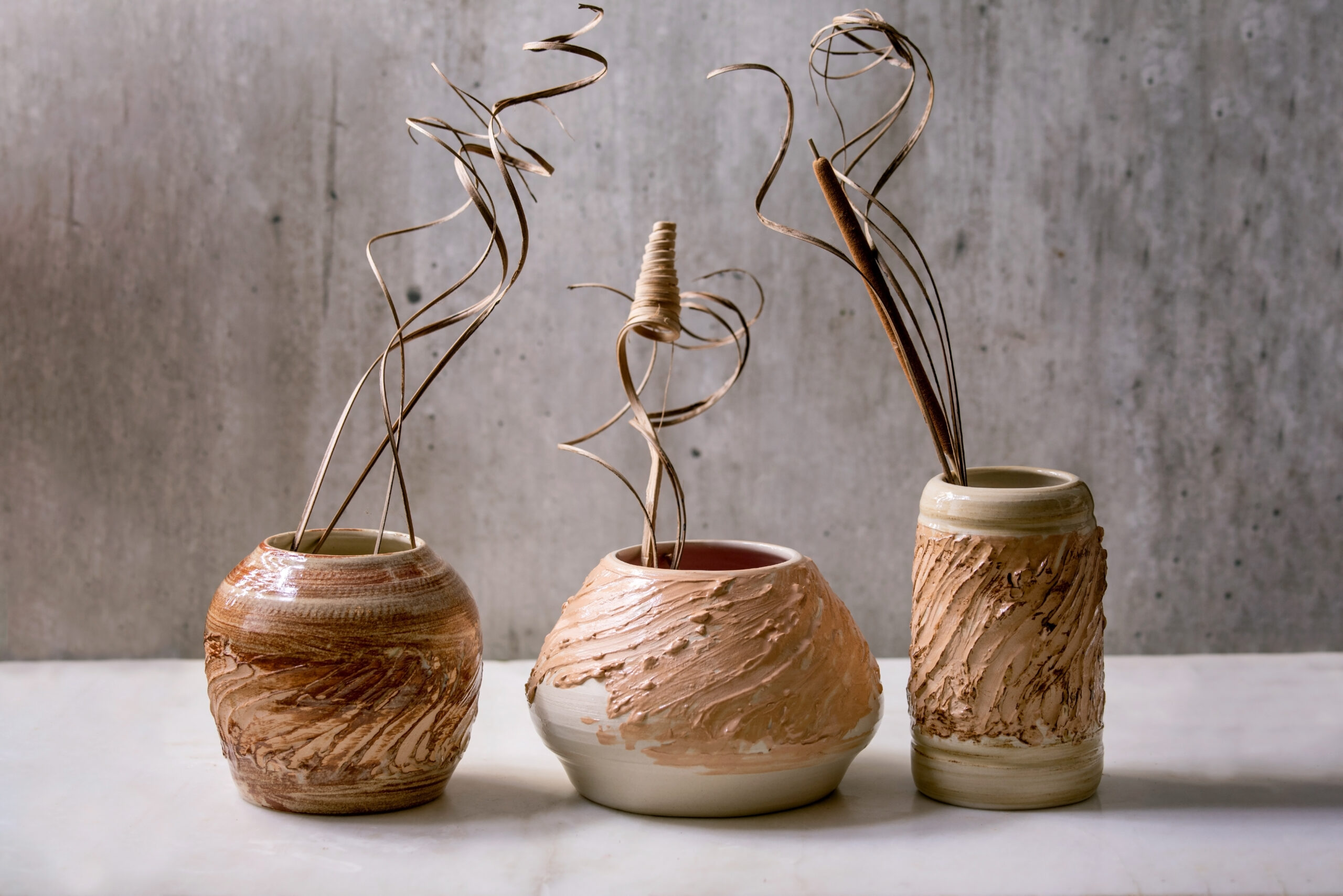 Three different shapes beige brown ceramic vases with dry flowers and twigs branch on white marble table with gray wall behind. Copy space.